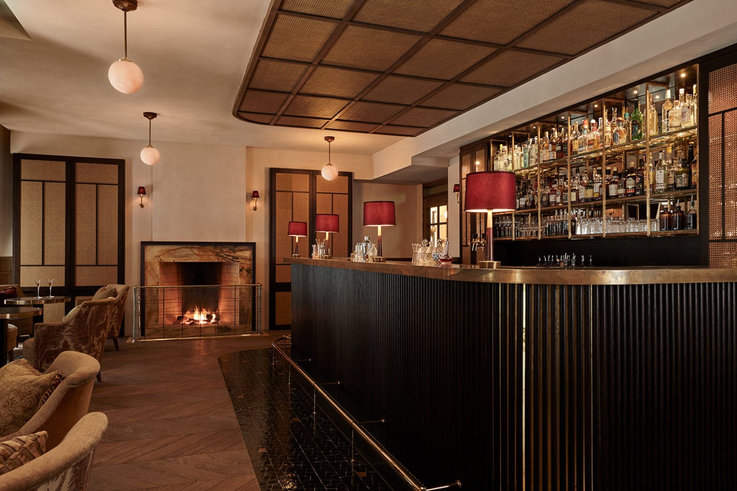 Hotel Sanders, Europe — Luxury bar lounge with brass-trimmed black counter, red-shaded pendant lights, fireplace, woven ceiling panels, and warm wood flooring.