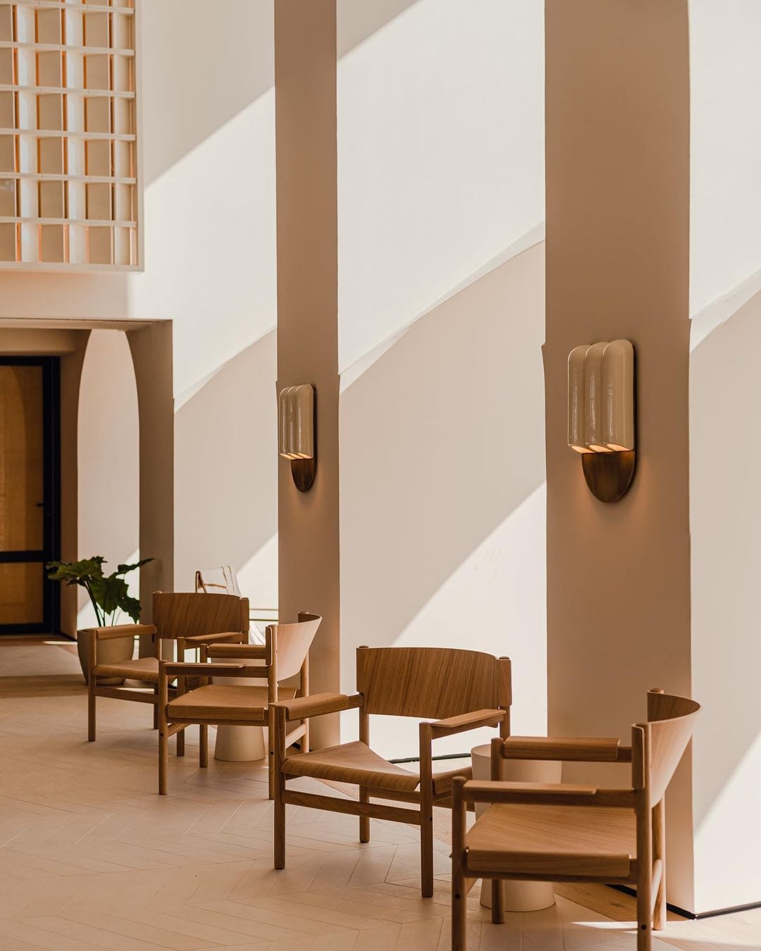Aethos Ericeira, Portugal — Minimalist corridor with timber chairs, brass wall sconces, and geometric sunlight casting shadows on whitewashed walls.