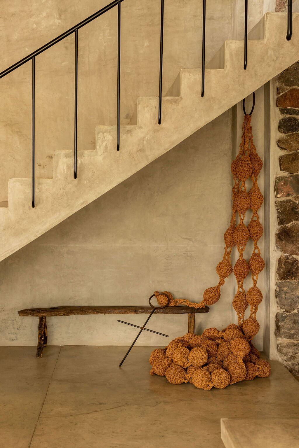 Villa ITO, Mexico — Minimalist stairwell with cream travertine walls, black metal railings, rustic wooden bench, and hanging terracotta spheres casting geometric shadows.
