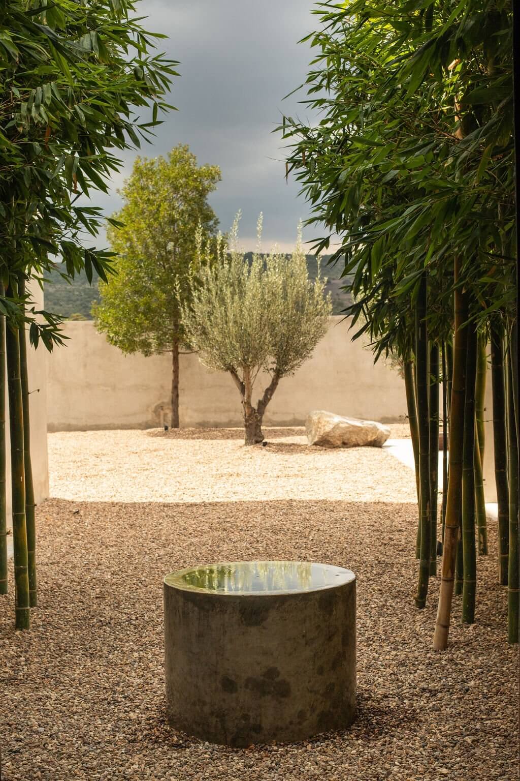 Villa ITO, Mexico — Minimalist courtyard with circular water basin, bamboo colonnade, and pruned trees on gravel, overcast sky and stone walls beyond.