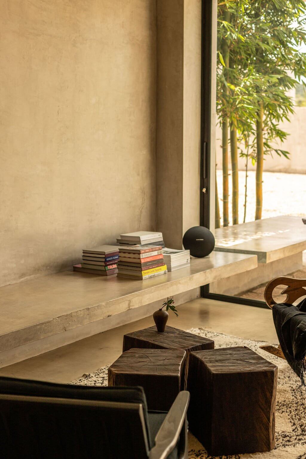 Villa ITO, Mexico — Minimalist living room with stone window seat, stacked books, black sphere speaker, wooden stools, and bamboo garden view through floor-to-ceiling glass doors.