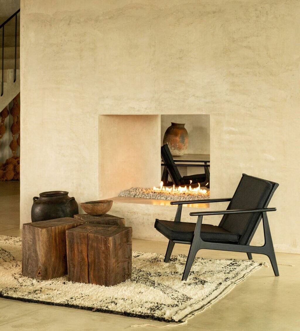 Villa ITO, Mexico — Minimalist lounge with mid-century modern chair, wooden stools, ceramic vessels, and built-in fireplace on textured cream interior.