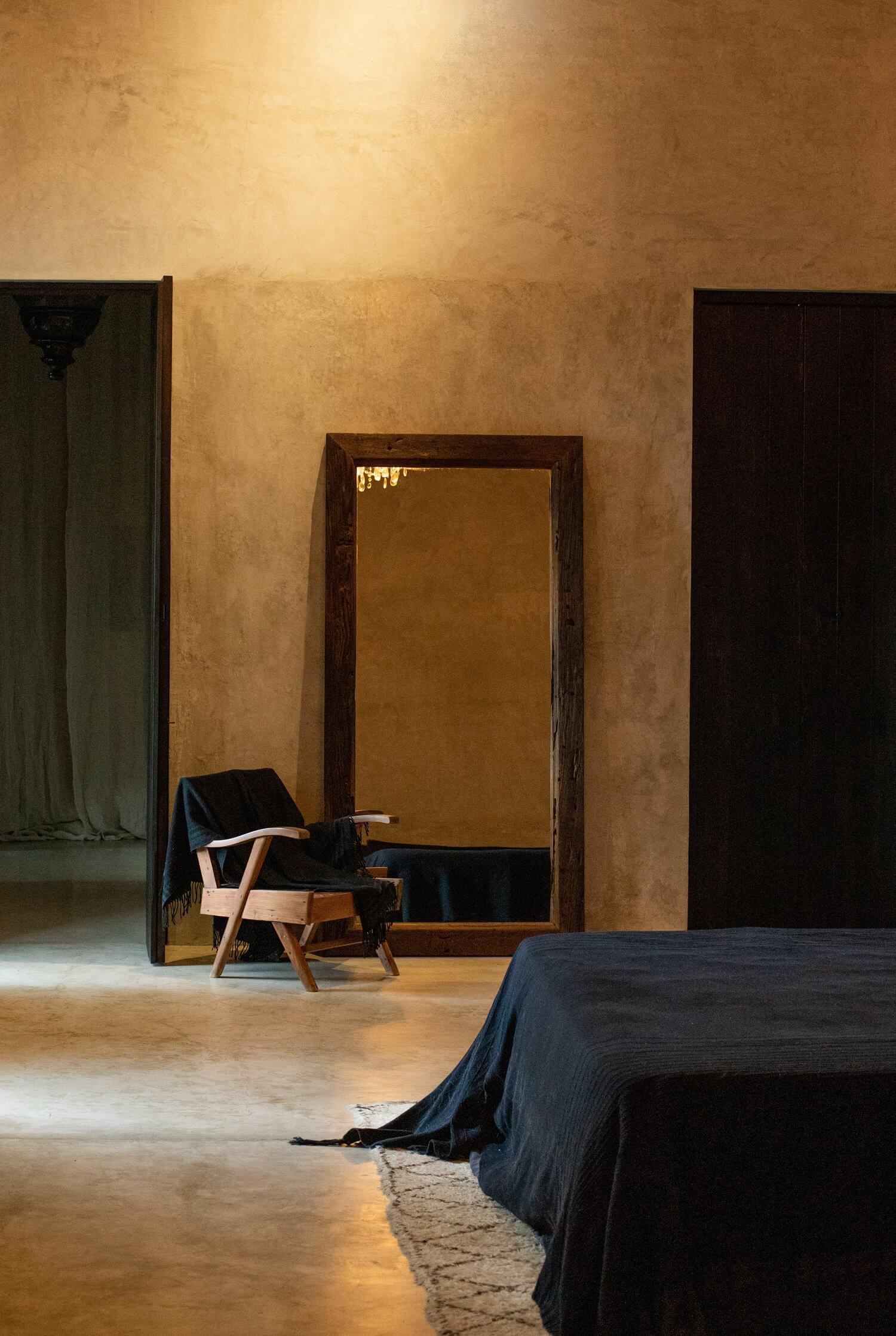Villa ITO, Mexico — Minimalist bedroom with ochre textured walls, wooden-framed mirror, mid-century chair with navy throw, polished concrete floors and natural light.