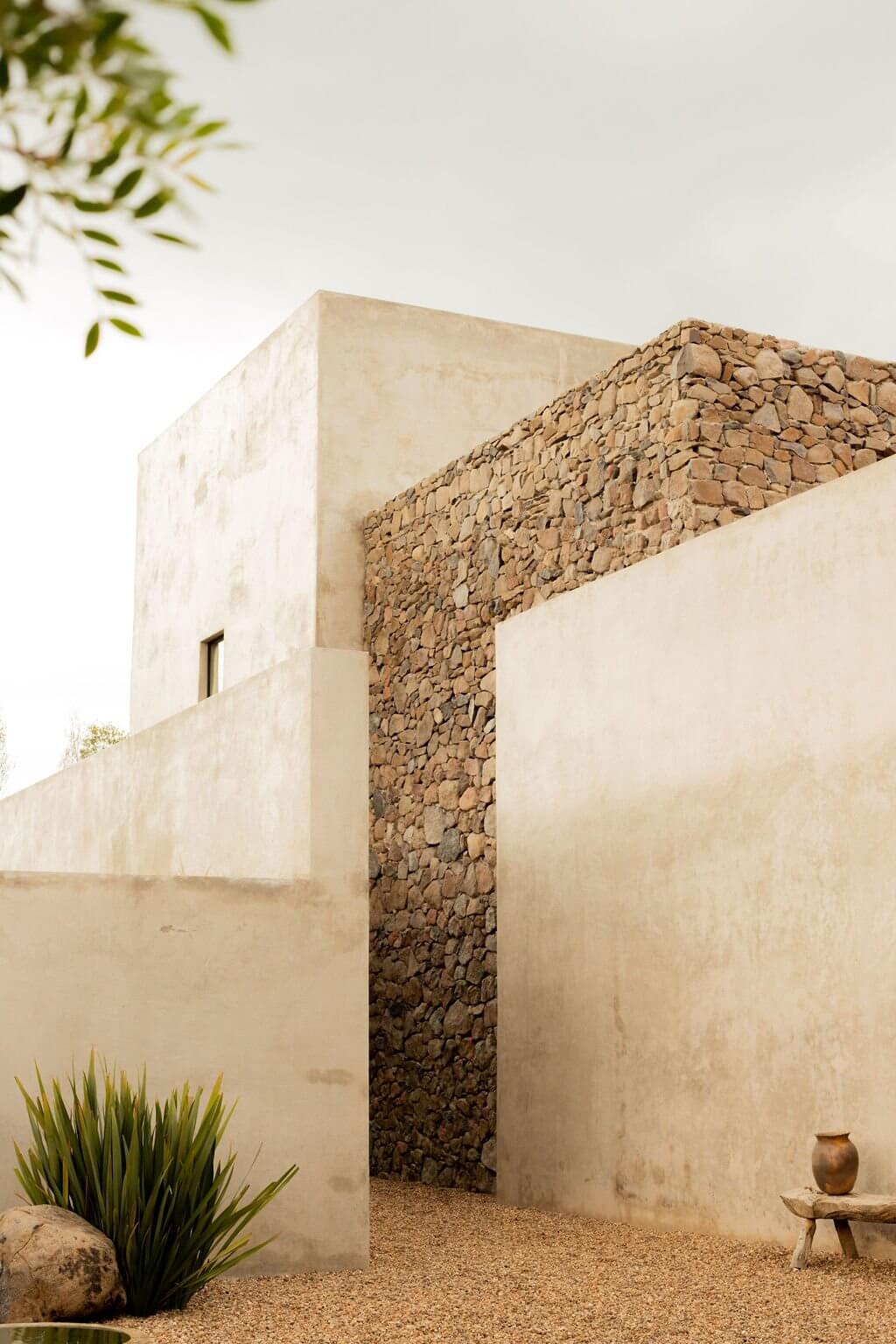 Villa ITO, Mexico — Minimalist stone and plaster courtyard with textured clay wall, small window detail, ceramic vessel, and desert landscaping under clear light.