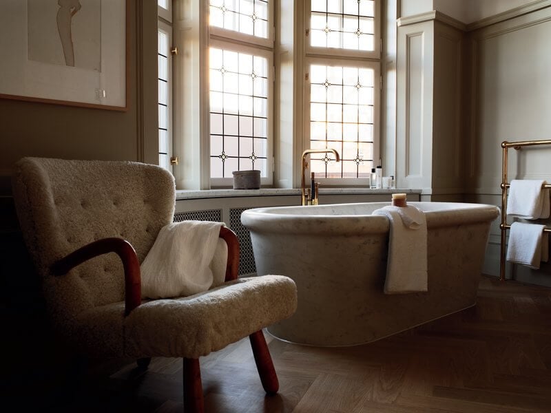 Ett Hem, Europe — Luxe bathroom with freestanding soaked tub, burgundy wingback chair, tall windows with geometric glazing, warm natural light, minimalist design.