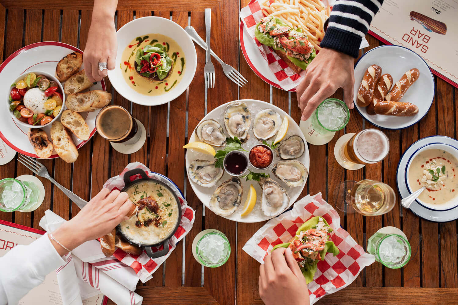 Sand Bar, North America — Overhead view of coastal dining table with oysters, seafood, fresh bread, and cocktails shared among guests on wooden deck.