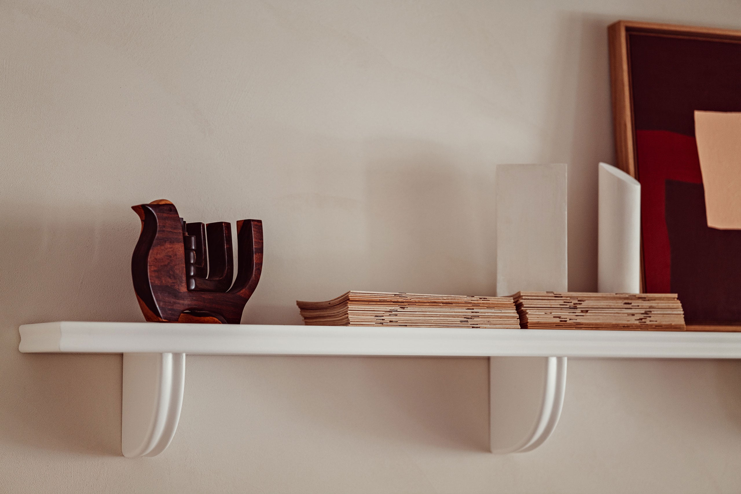 Académies et des Arts, Europe — White floating shelf displaying dark wood sculptural object, stacked papers, and framed artworks against neutral wall.