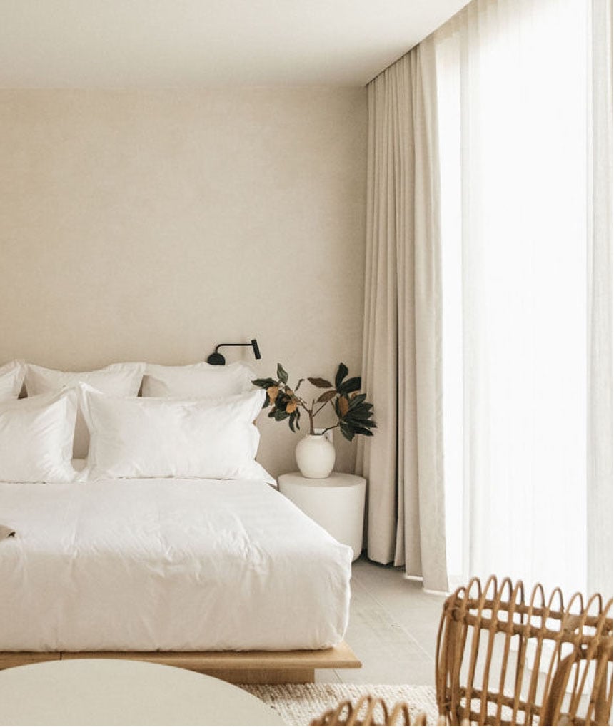 Octavia Casa, Mexico — Minimalist bedroom with white linens, cream walls, floor-to-ceiling curtains, woven chair, and fresh flowers in ceramic vase by nightstand.
