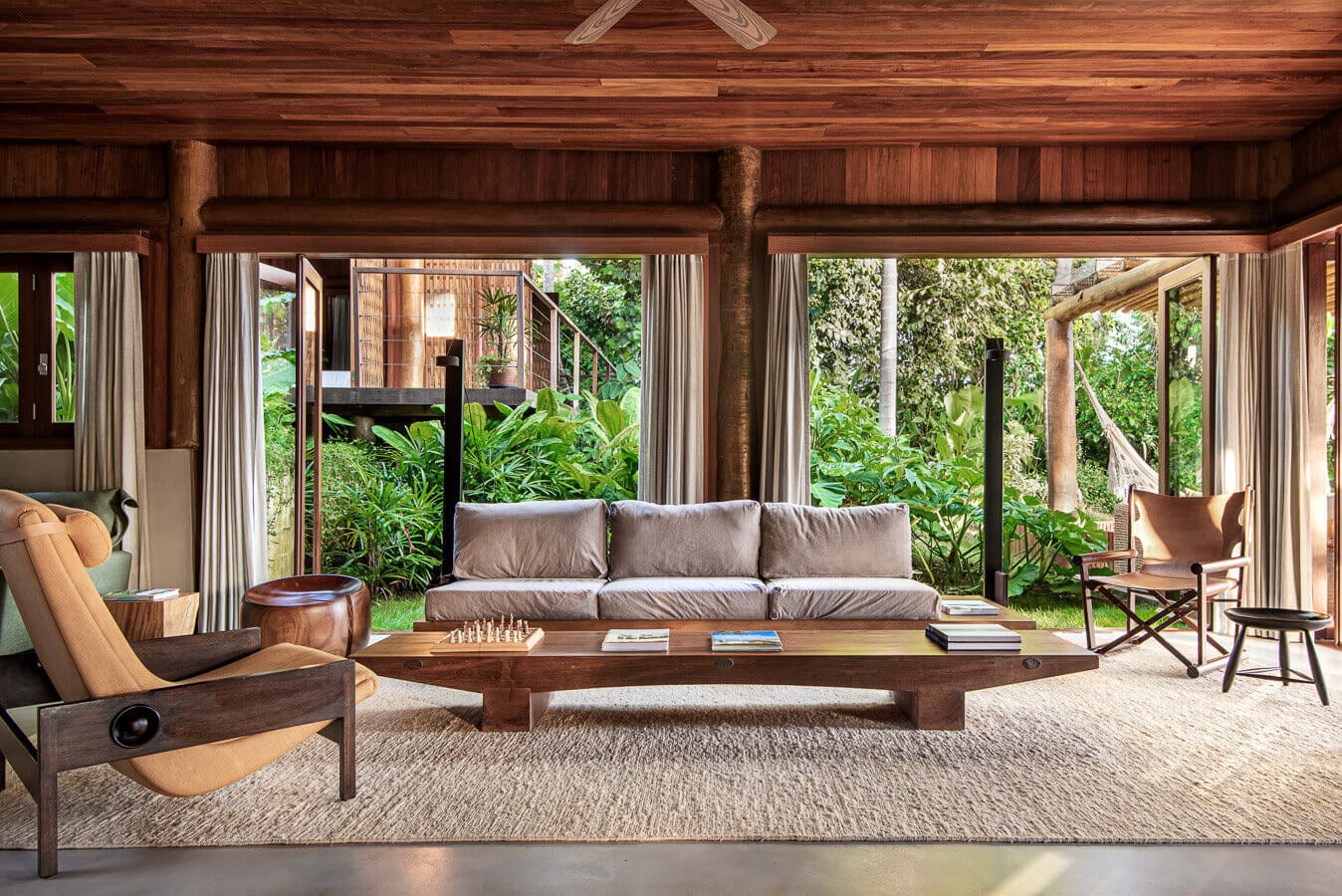 Barracuda Hotel & Villas, South America — Teak-beamed living room with linen sofa, leather chairs, wooden coffee table, and floor-to-ceiling glass doors opening to tropical gardens.