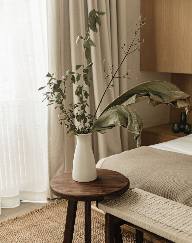 Octavia Casa, Mexico — Minimalist bedside arrangement with dried botanicals in cream ceramic vase on dark wood round table, natural linen textiles, warm neutral tones.
