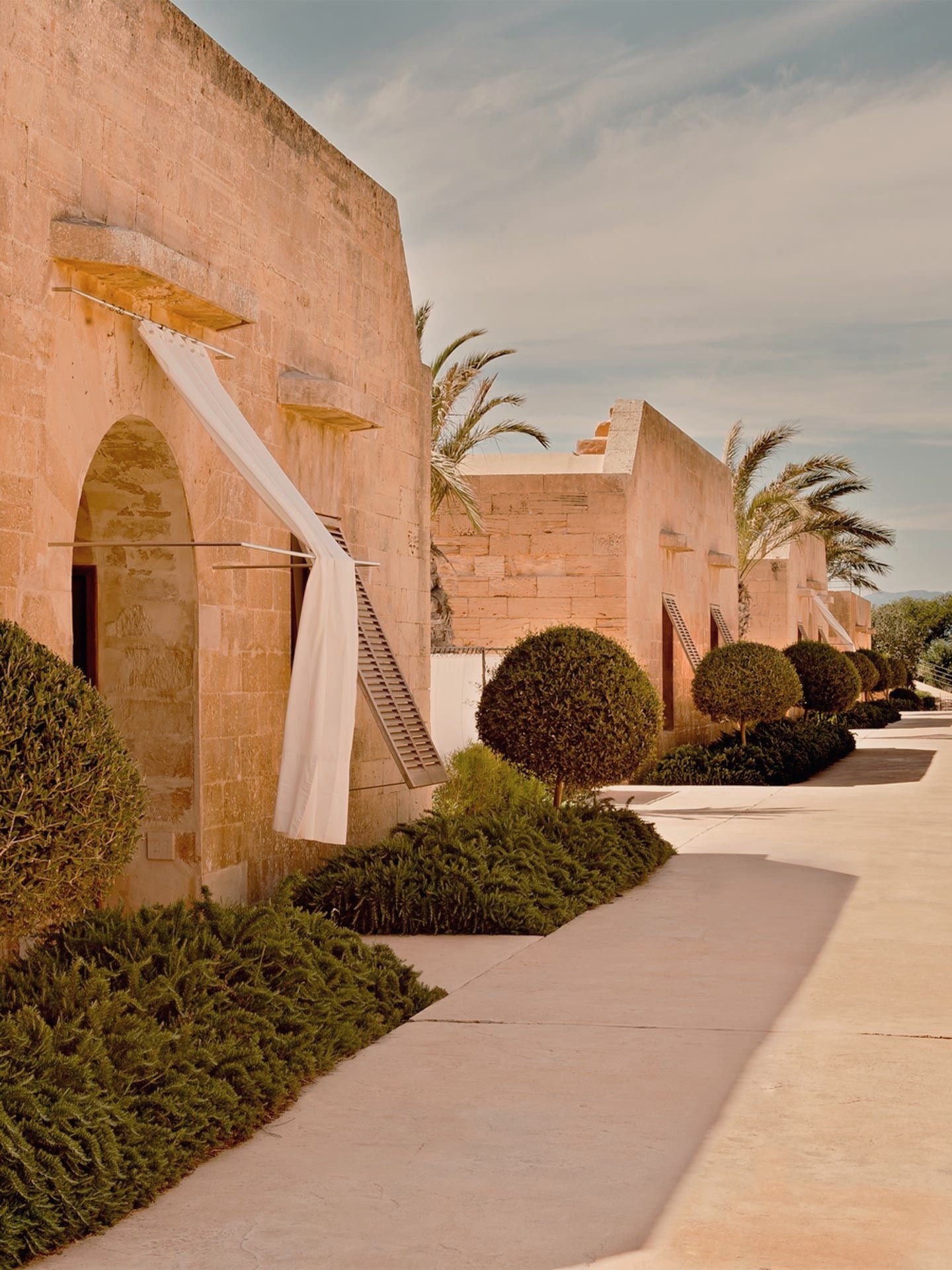 Cap Rocat, Balearic Islands, Spain — Terracotta fortress with arched doorways, geometric topiary, palm trees, and flowing white fabric in Mediterranean sunlight.