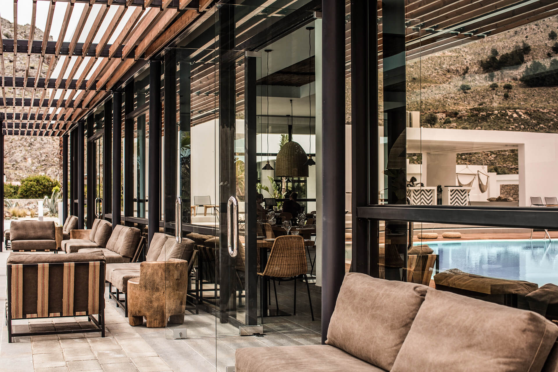 Casa Cook Rhodes, Greece — Modern poolside pavilion with wooden pergola, black steel columns, floor-to-ceiling glass doors, neutral upholstered seating, and stone accent walls overlooking Mediterranean landscape.