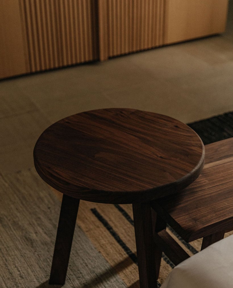 Octavia Casa, Mexico — Dark wood stool with round seat beside bench in minimalist interior with warm lighting and corrugated wall panels.