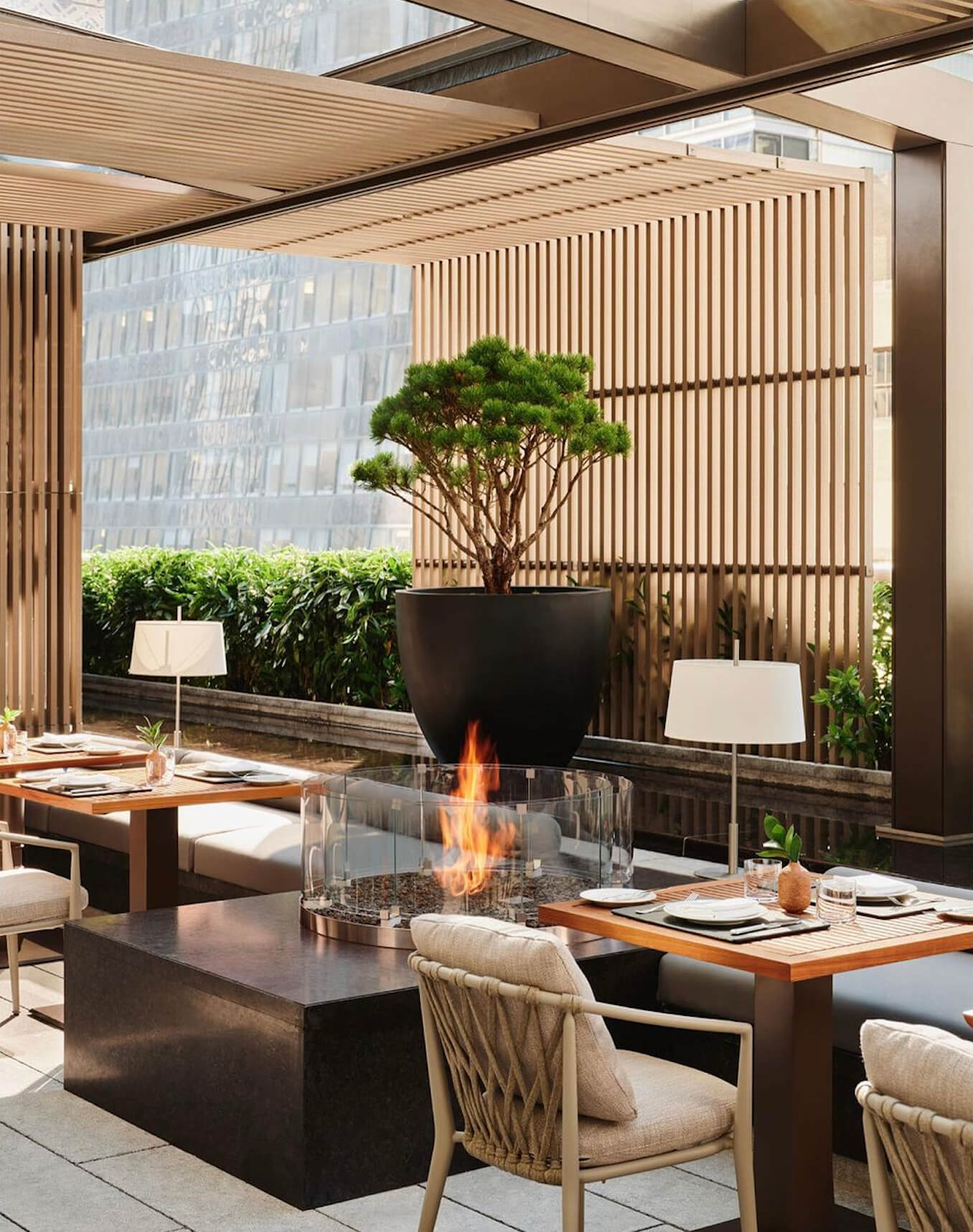 Aman New York, New York, United States — Modern rooftop terrace with wooden slatted screens, dining tables, tabletop fireplace, sculptural tree, and city skyline views.