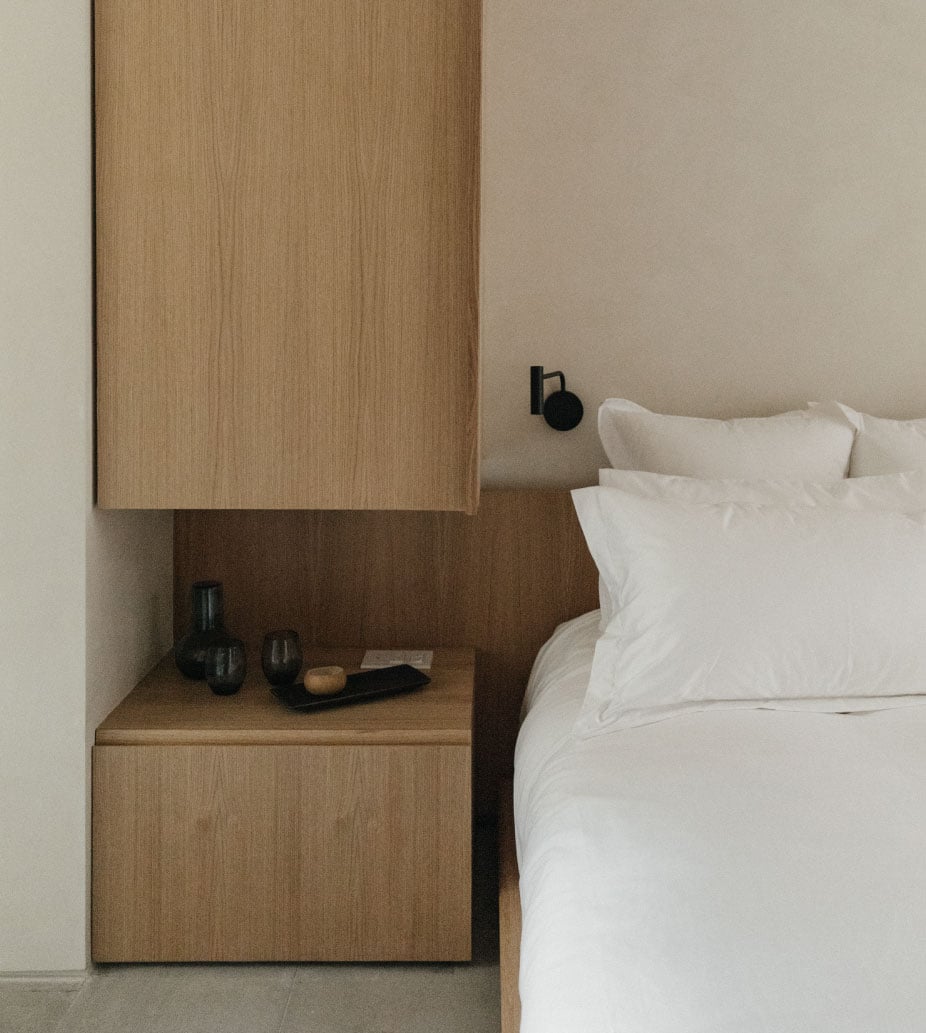 Octavia Casa, Mexico — Minimalist bedroom with natural wood nightstand, white linens, and black sconce lighting against neutral walls.