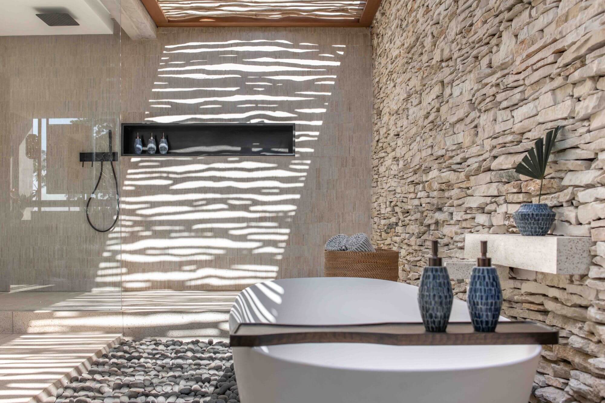 Cap Karoso, Asia — Luxury spa bathroom with sculptural white soaking tub, stacked stone accent wall, geometric skylight casting striped shadows, and minimalist blue ceramic vessels.