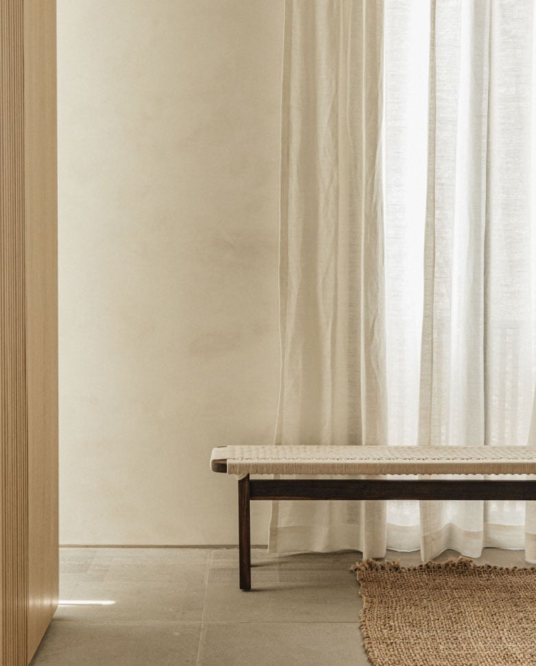 Octavia Casa, Mexico — Minimalist bedroom corner with wooden bench, linen curtains, woven rug, and soft natural light filtering through sheer drapes against neutral plaster walls.