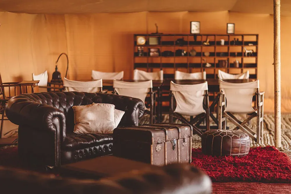 Scarabeo Camp, Africa — Leather chesterfield sofa with canvas chairs and vintage luggage on red kilim rug in warm-lit glamping tent interior.