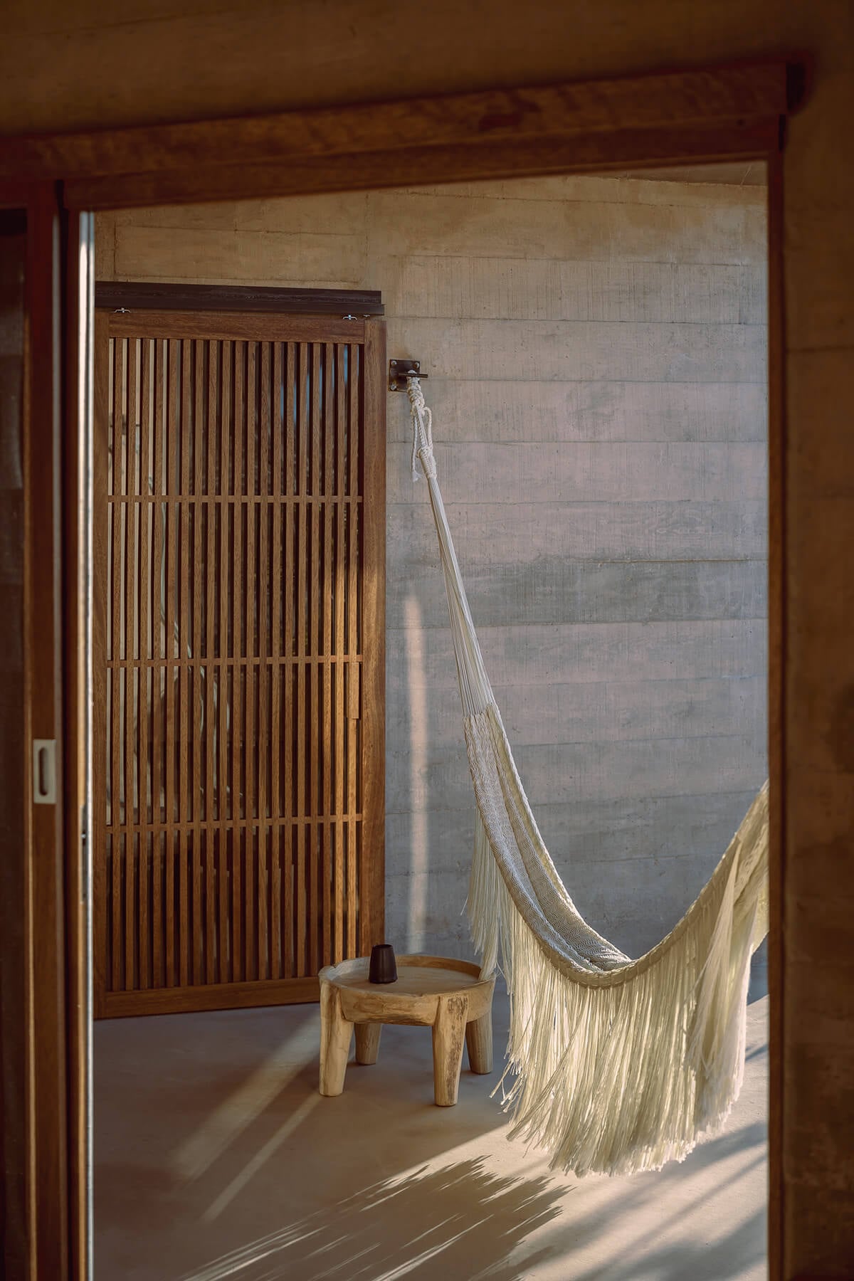 Paradero Todos Santos, Mexico — Minimalist bedroom with wooden slatted door, woven hammock, concrete walls, and natural light casting geometric shadows