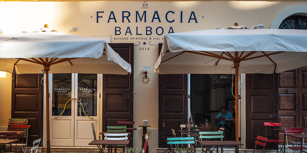 Farmacia Balboa's cream-and-timber facade glows in afternoon light, stacked colorful chairs nestled beneath canvas umbrellas.