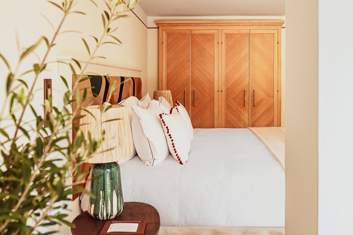 Hôtel Lou Pinet, Europe — Sunlit bedroom with white linens, cognac wood wardrobe, potted plant, and warm neutral palette.