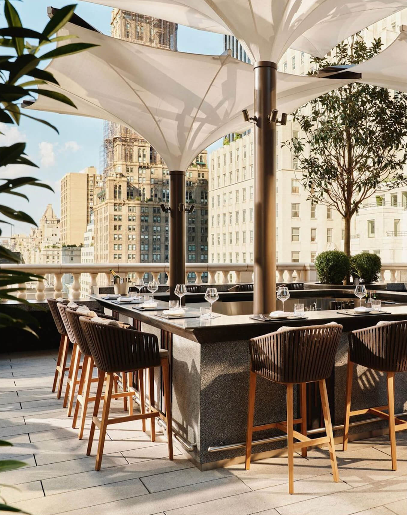 Aman New York, New York, United States — Rooftop dining terrace with cream sails, wooden bar seating, Manhattan skyline views, and sculptural landscaping.