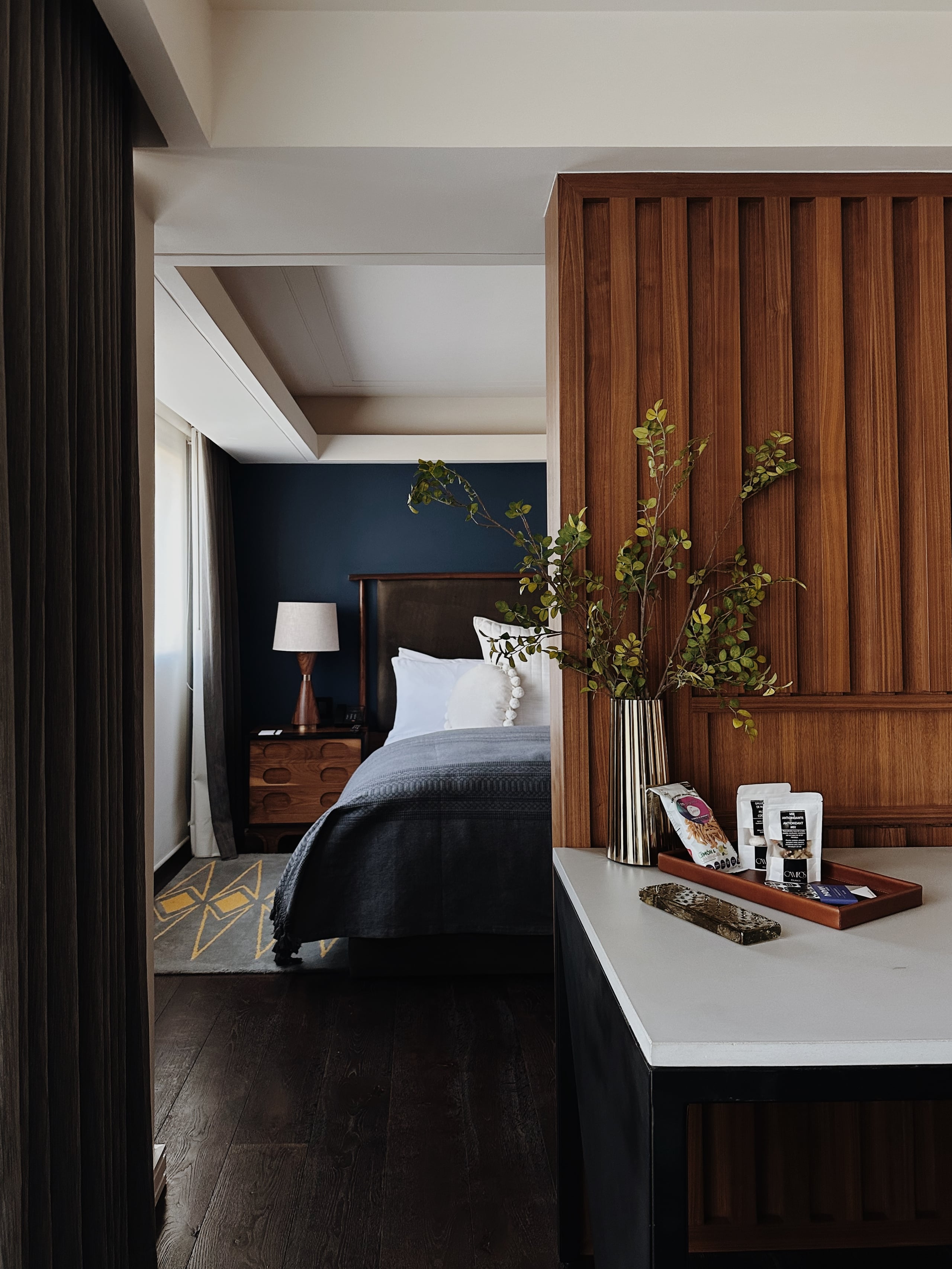 Campos Polanco, Mexico — Modern bedroom suite with walnut wood paneling, navy accent wall, dark hardwood floors, and minimalist desk workspace with fresh greenery arrangement.