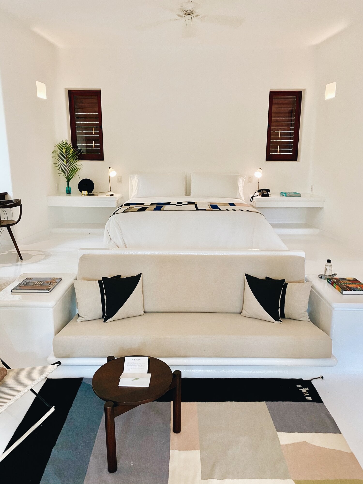 Hotel Esencia, Mexico — Minimalist bedroom with cream linen sofa, dark wood shutters, floating nightstands, geometric area rug, and mid-century side table in bright natural light.