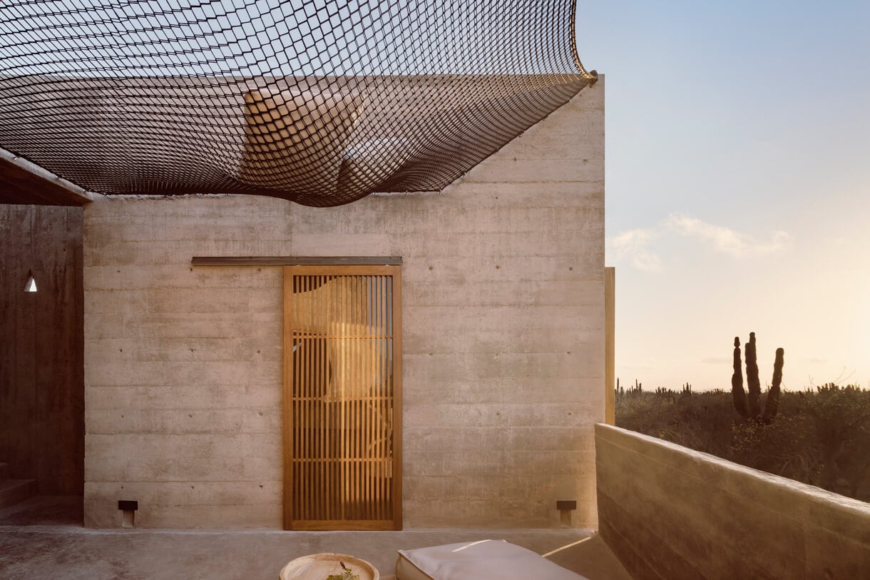 Paradero Todos Santos, Mexico — Minimalist concrete terrace with wooden slatted door, mesh canopy, and desert landscape at sunset.