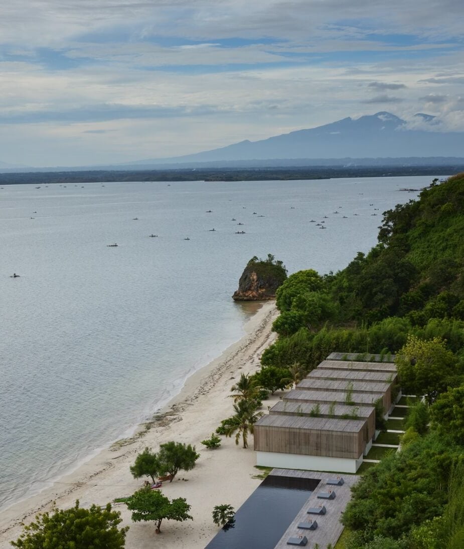Innit Lombok, Indonesia — Wooden pavilions on white sand beach with Mount Agung view, infinity pool, and traditional outrigger boats dotting turquoise waters.