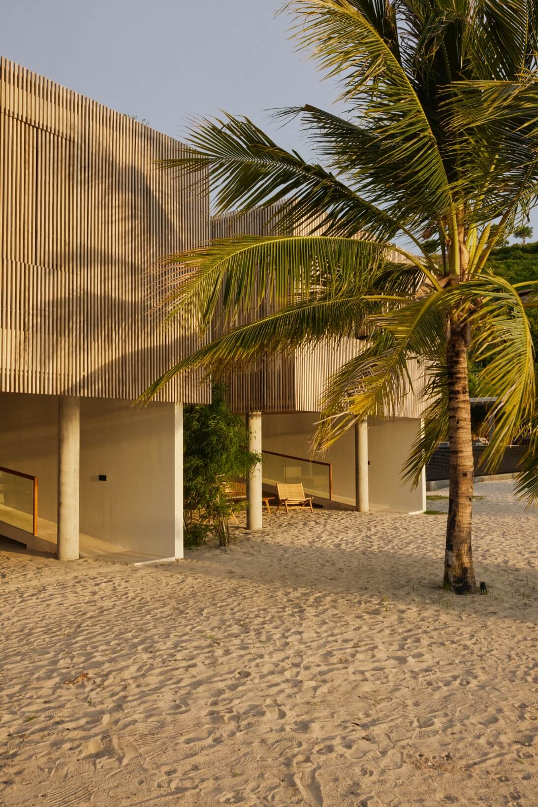 Innit Lombok, Indonesia — Modernist beachfront pavilion with corrugated timber screening, concrete pillars, and palm trees casting shadows on sand.