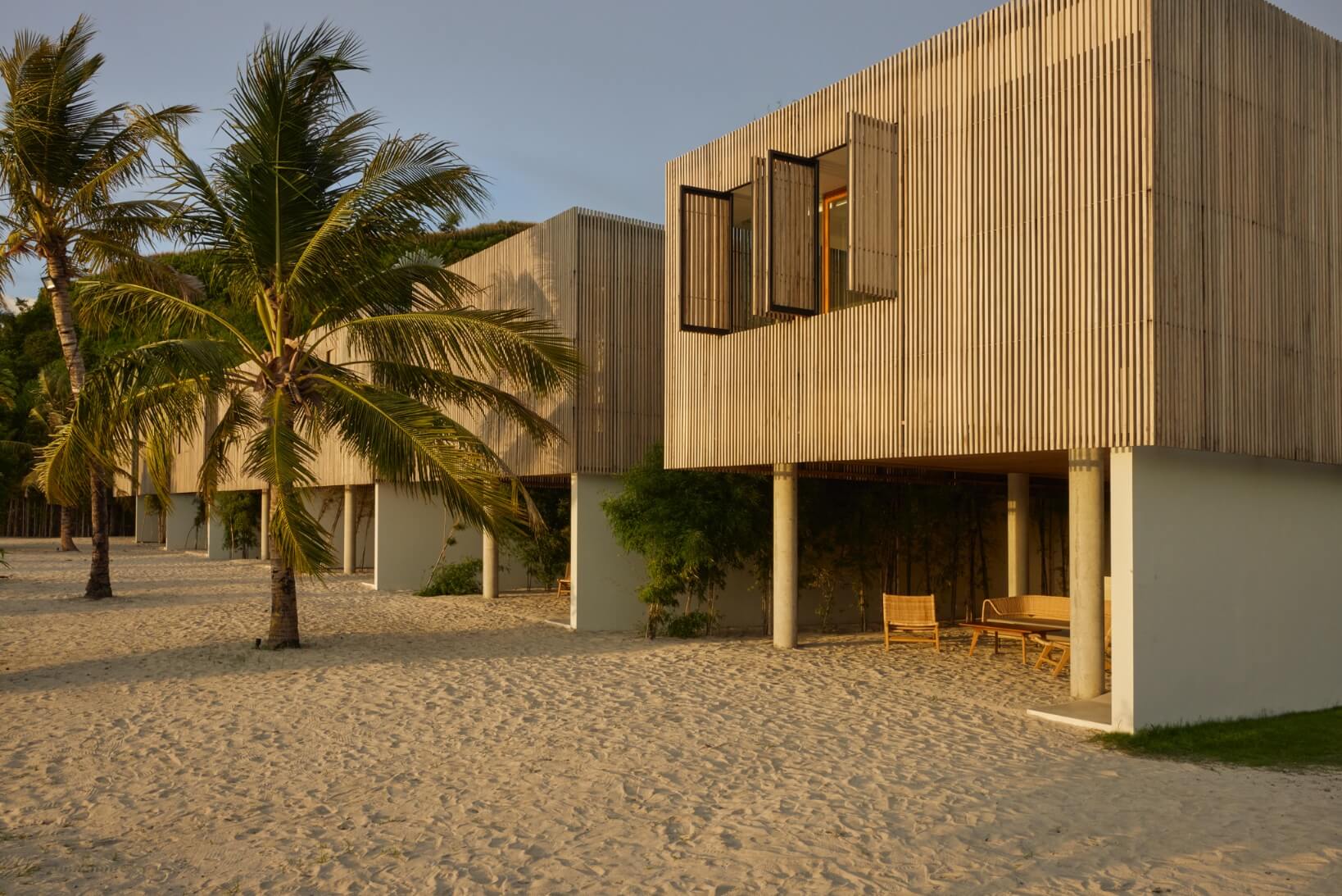 Innit Lombok, Asia — Minimalist beach villa with timber-slatted facade, concrete pillars, and open-air terrace on sand, palm trees, golden hour light.