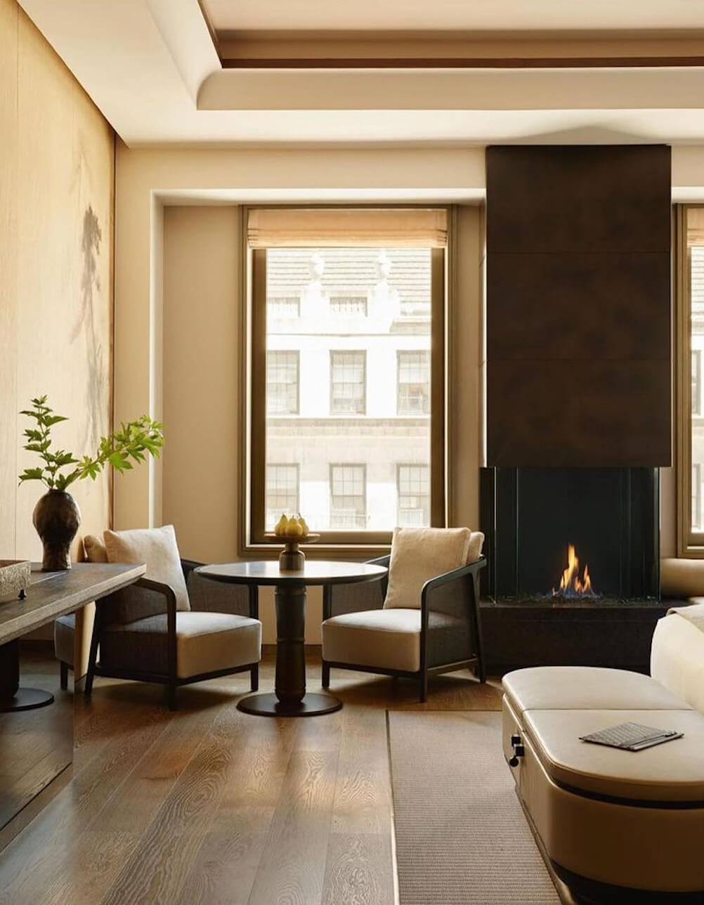 Aman New York, New York, United States — Modern living room with cream upholstered chairs, black fireplace, floor-to-ceiling windows overlooking city, natural wood flooring, minimalist luxury interior design.