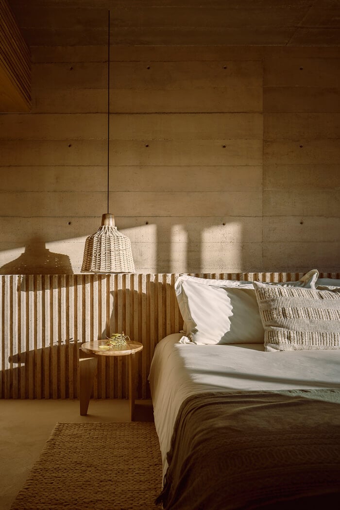 Paradero Todos Santos, Mexico — Minimalist bedroom with concrete walls, wooden slat partition, woven pendant light, and natural sunlight casting geometric shadows across neutral linens and textures.