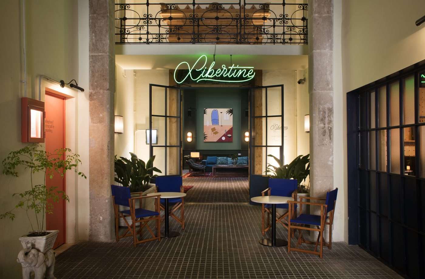 Casa Bonay, Barcelona, Spain — Neon-lit gallery entrance with black steel-frame doors, blue mid-century chairs, warm ambient lighting, and ornate wrought-iron balcony above.
