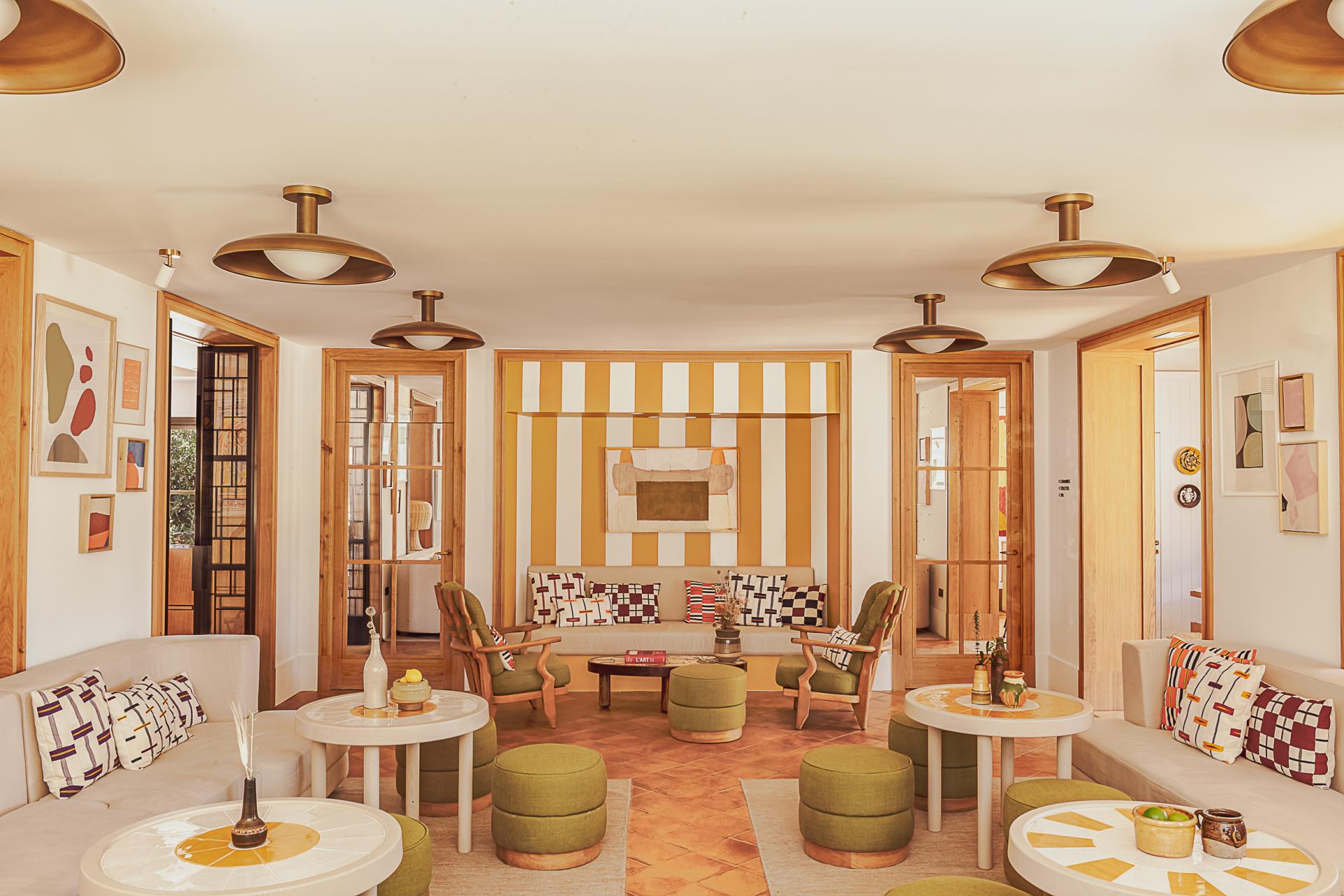 Hôtel Lou Pinet, Europe — Mid-century modern lounge with golden wood furnishings, striped accent wall, brass pendant lights, and geometric patterned textiles in warm earth tones.