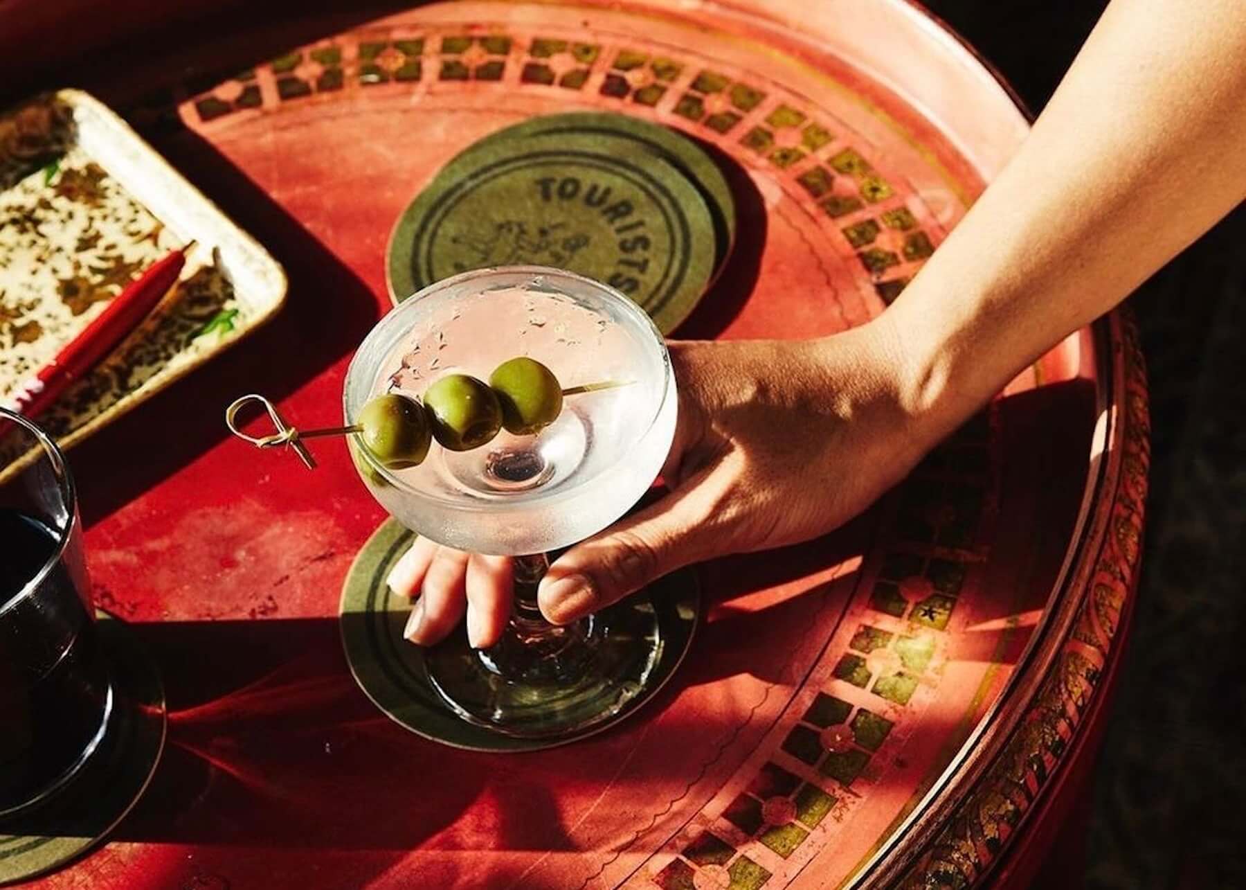 Tourists Hotel — hand holding olive-garnished cocktail over red velvet roulette table with brass details, luxury hospitality ambiance.