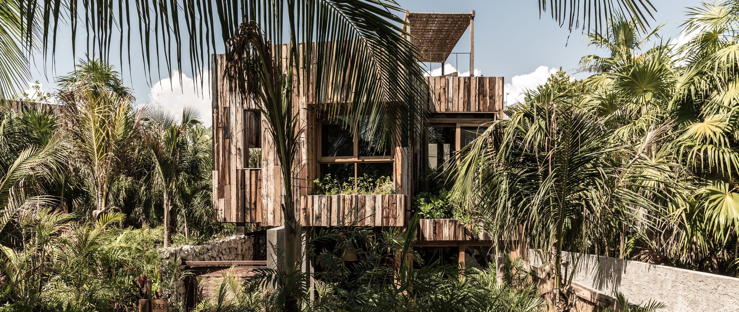 Be Tulum, Mexico — Weathered timber structure with stacked levels and open balconies nestled among lush palm trees under bright sky.