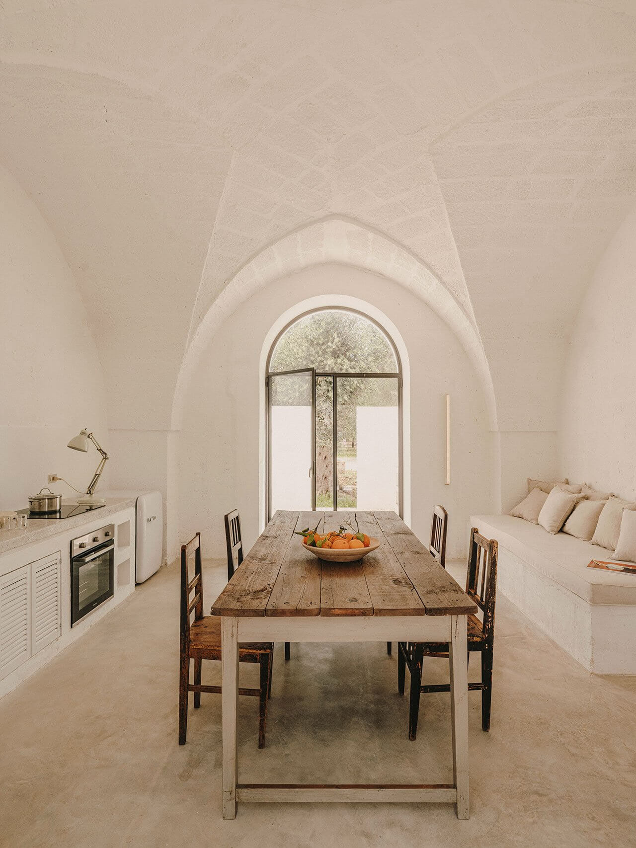 Borgo Gallana, Europe — Barrel-vaulted dining room with arched window, rustic wooden table, cream pillows on built-in seating, minimalist kitchen, natural light.