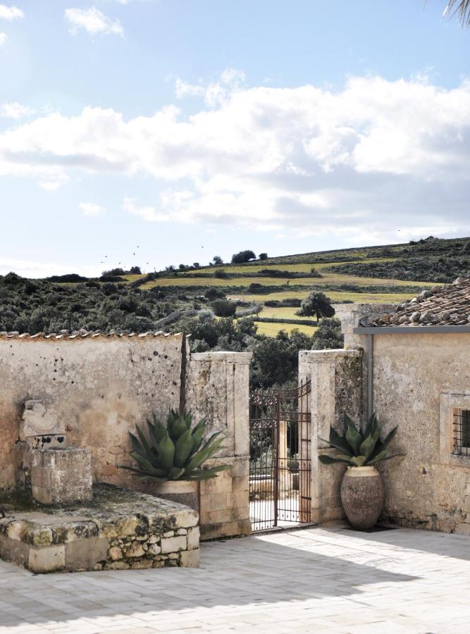 Dimora delle Balze, Europe — Weathered stone courtyard with agave plants in terracotta vessels, iron gate, and rolling Tuscan hills under blue sky.