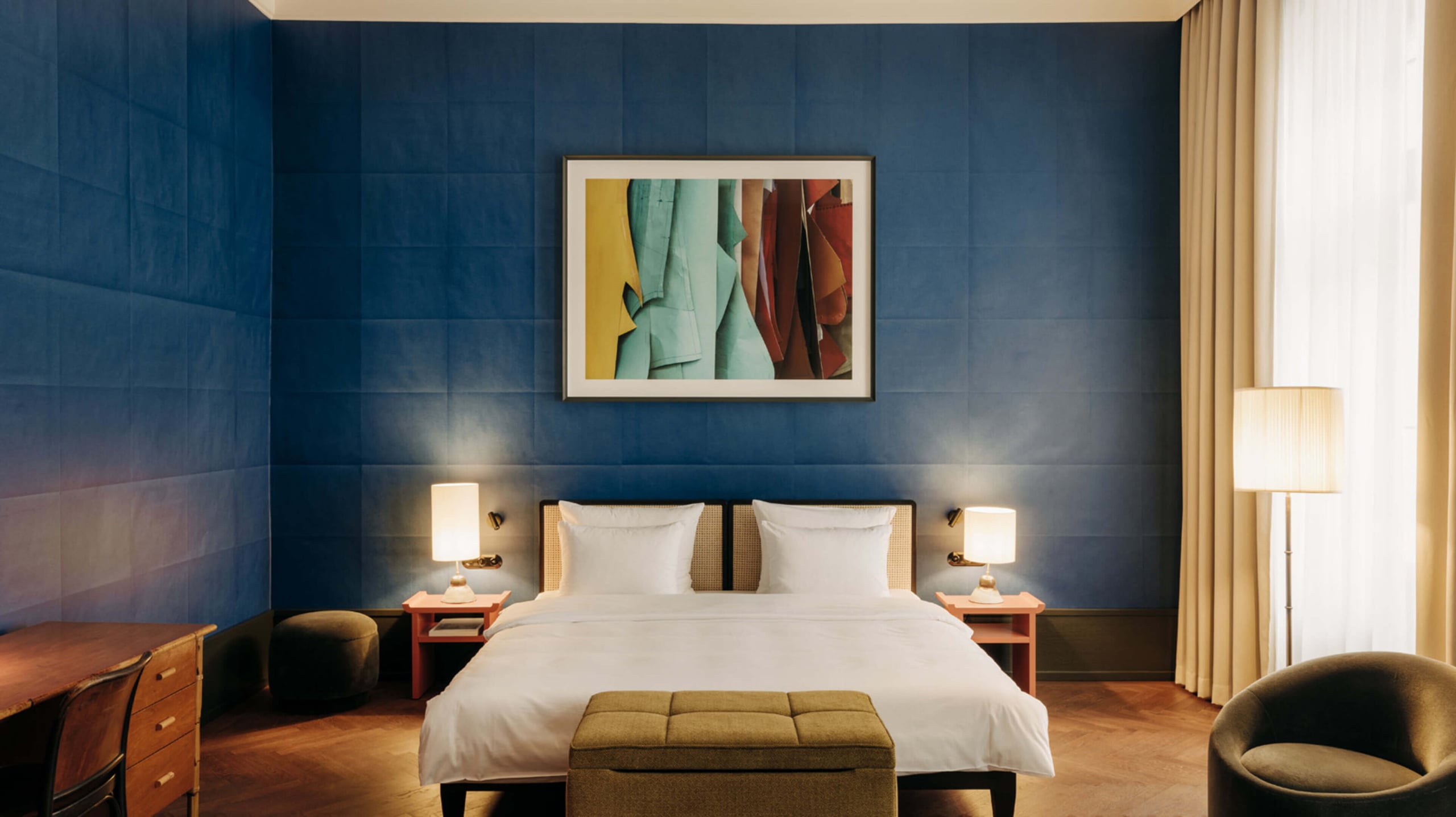 Château Royal, Europe — Contemporary hotel bedroom with deep blue paneled walls, white bedding, rattan headboard details, warm wood flooring, and gallery artwork above bed.