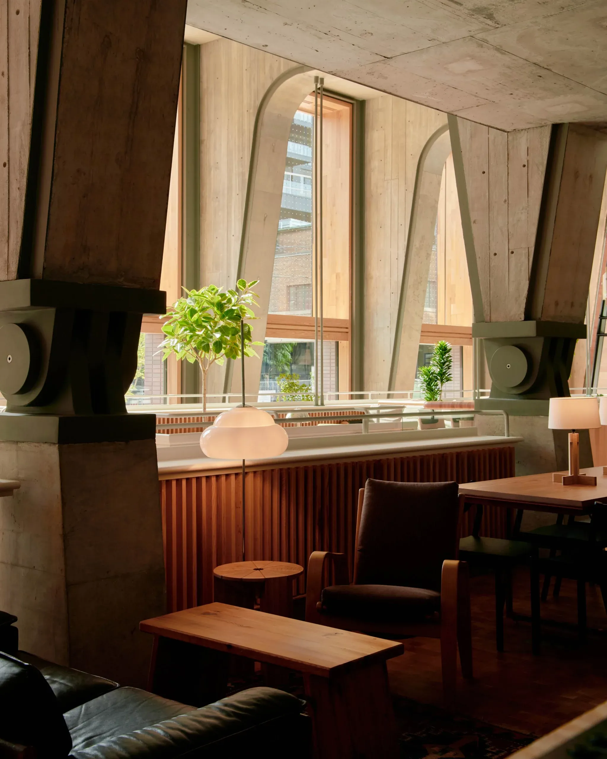 Ace Hotel Toronto, Canada — Modernist lounge with curved mirror, wood furnishings, potted plants, and floor-to-ceiling windows overlooking brick cityscape.