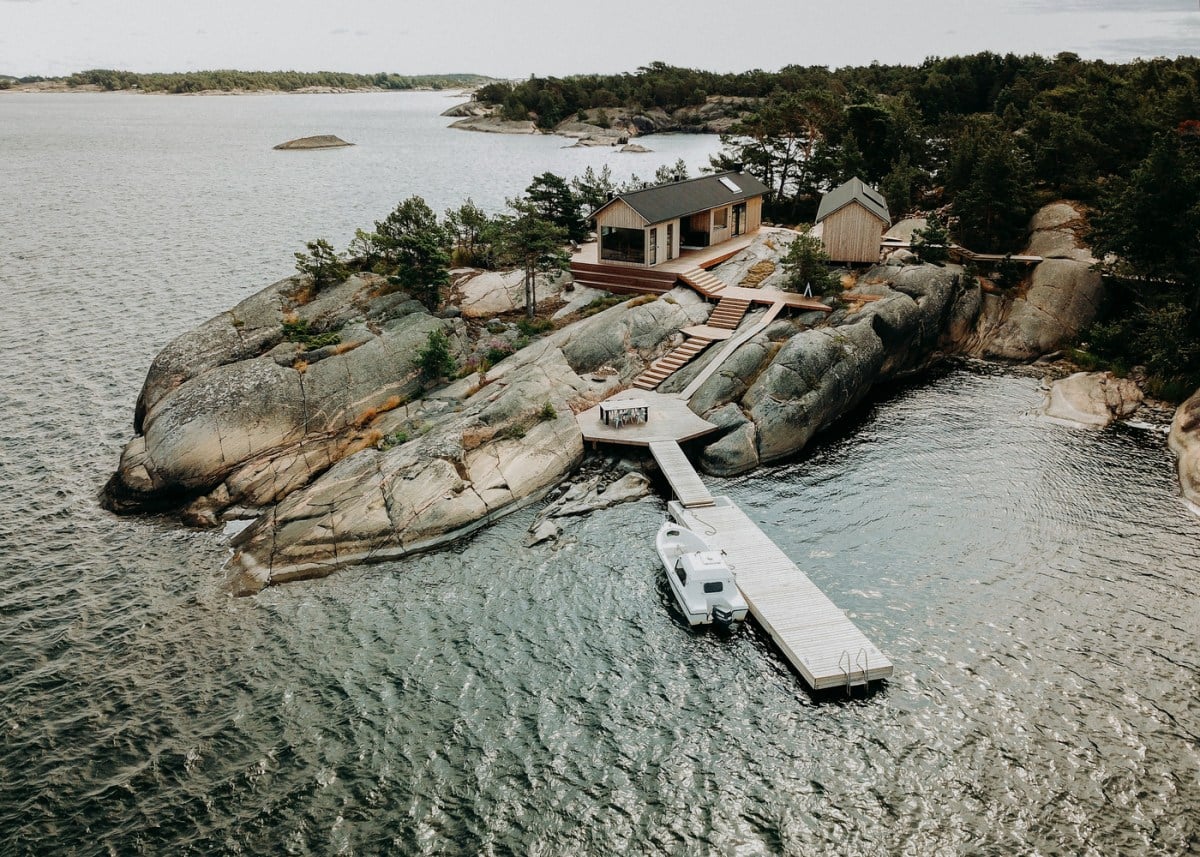 Project Ö — Modern wooden cabin with dark roof on rocky granite island, white dock with boat, surrounded by calm water and forested shoreline.