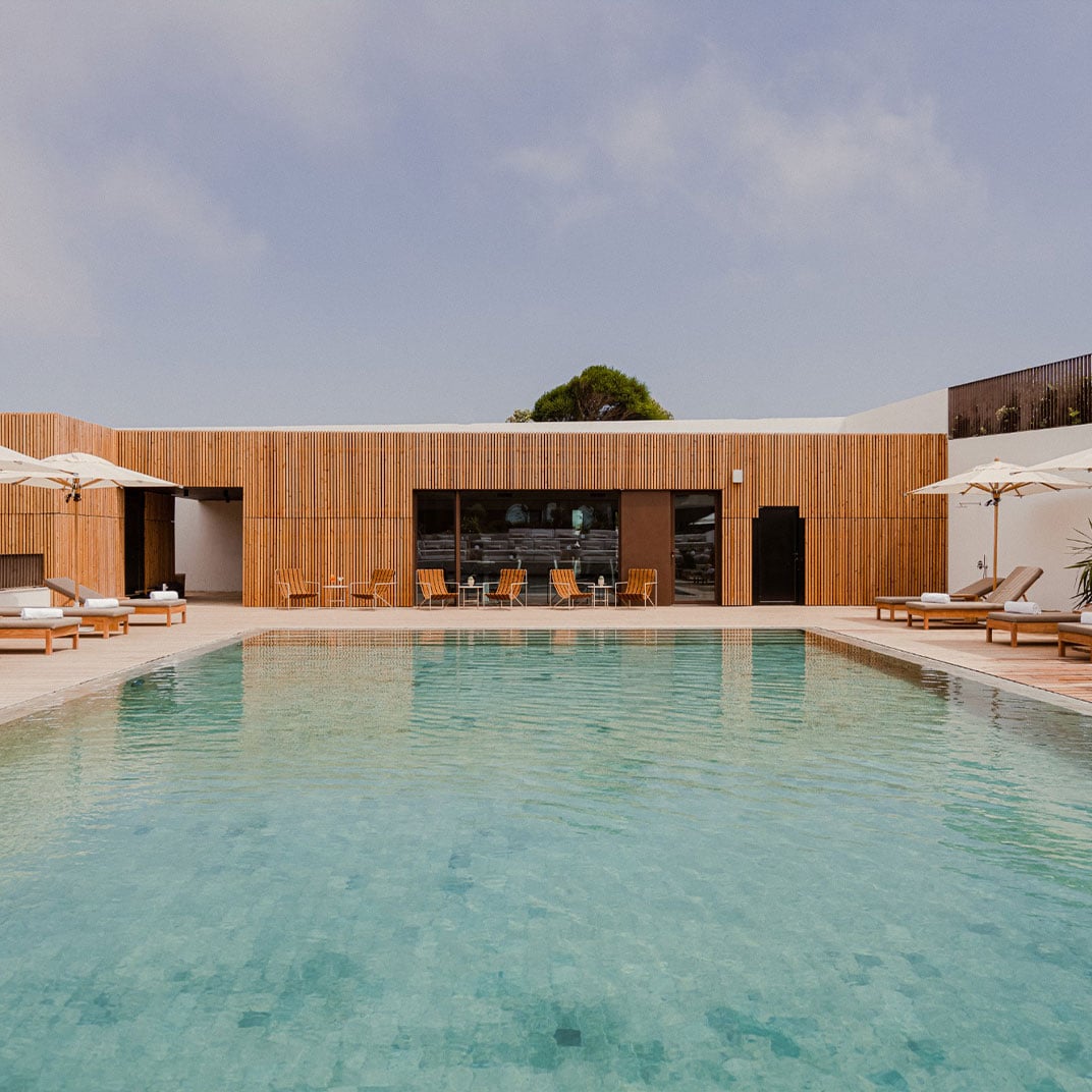 Aethos Ericeira, Portugal — Modern wooden-clad poolhouse with turquoise infinity pool, teak loungers, and minimalist coastal architecture under clear sky.