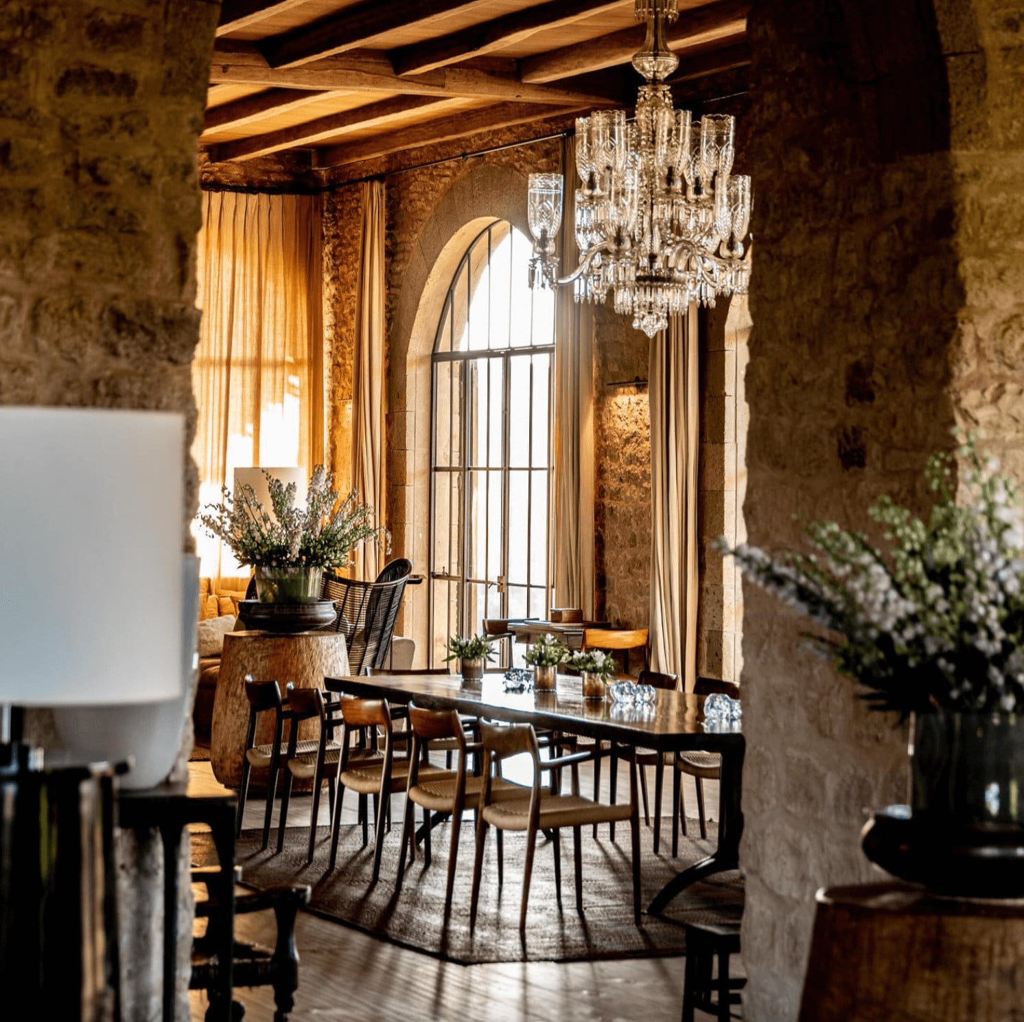 Arijiju Retreat, Africa — Dining room with exposed stone walls, wooden beams, crystal chandelier, arched windows with golden curtains, and black chairs around dark table.