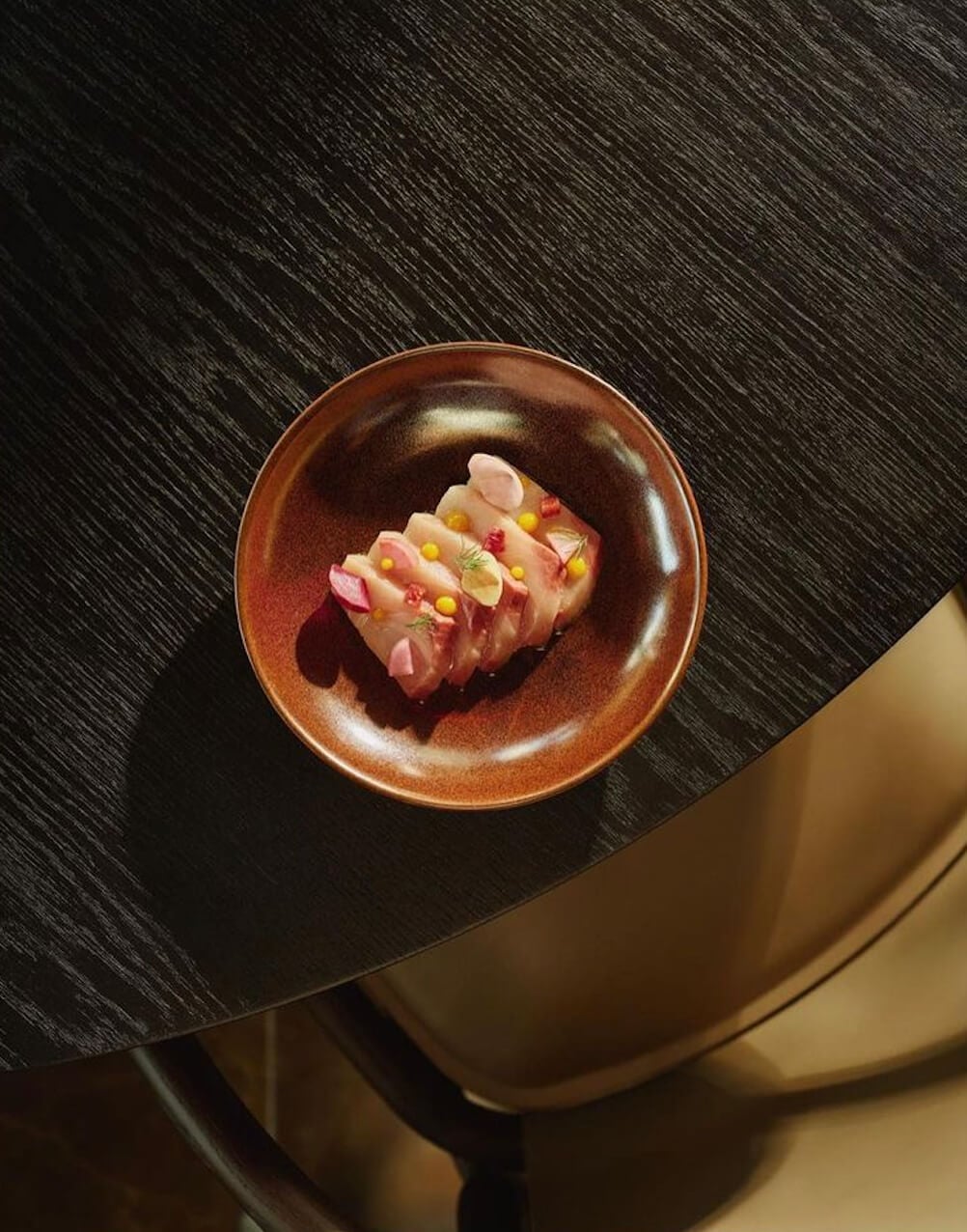 Aman New York, New York, United States — Pink scallop crudo with corn kernels and petals on rust ceramic bowl, minimalist plating on dark wood surface.