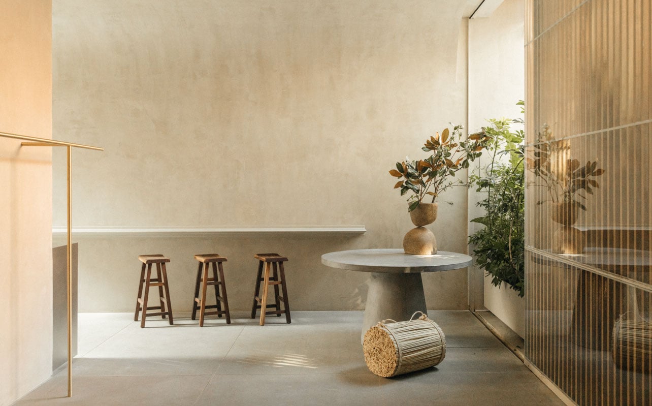 Octavia Casa, Mexico — Minimalist courtyard with natural light, concrete pedestal table, wooden bar stools, potted plants, and corrugated metal screening.