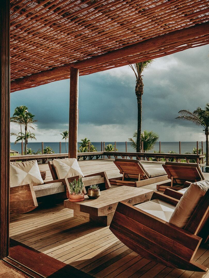 Barracuda Hotel & Villas, South America — Oceanfront terrace with teak lounge chairs, woven pergola, palm trees, and dramatic clouded sky over sea.
