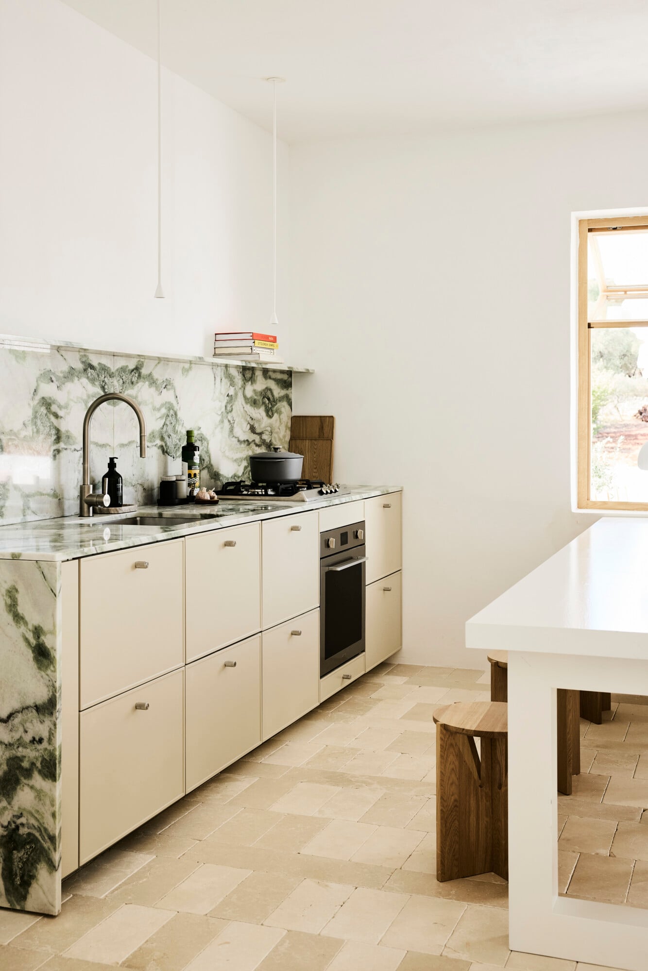Villa Sessana, Italy — Minimalist kitchen with cream cabinetry, veined marble backsplash and countertop, black fixtures, wooden stools, and natural light from large windows.