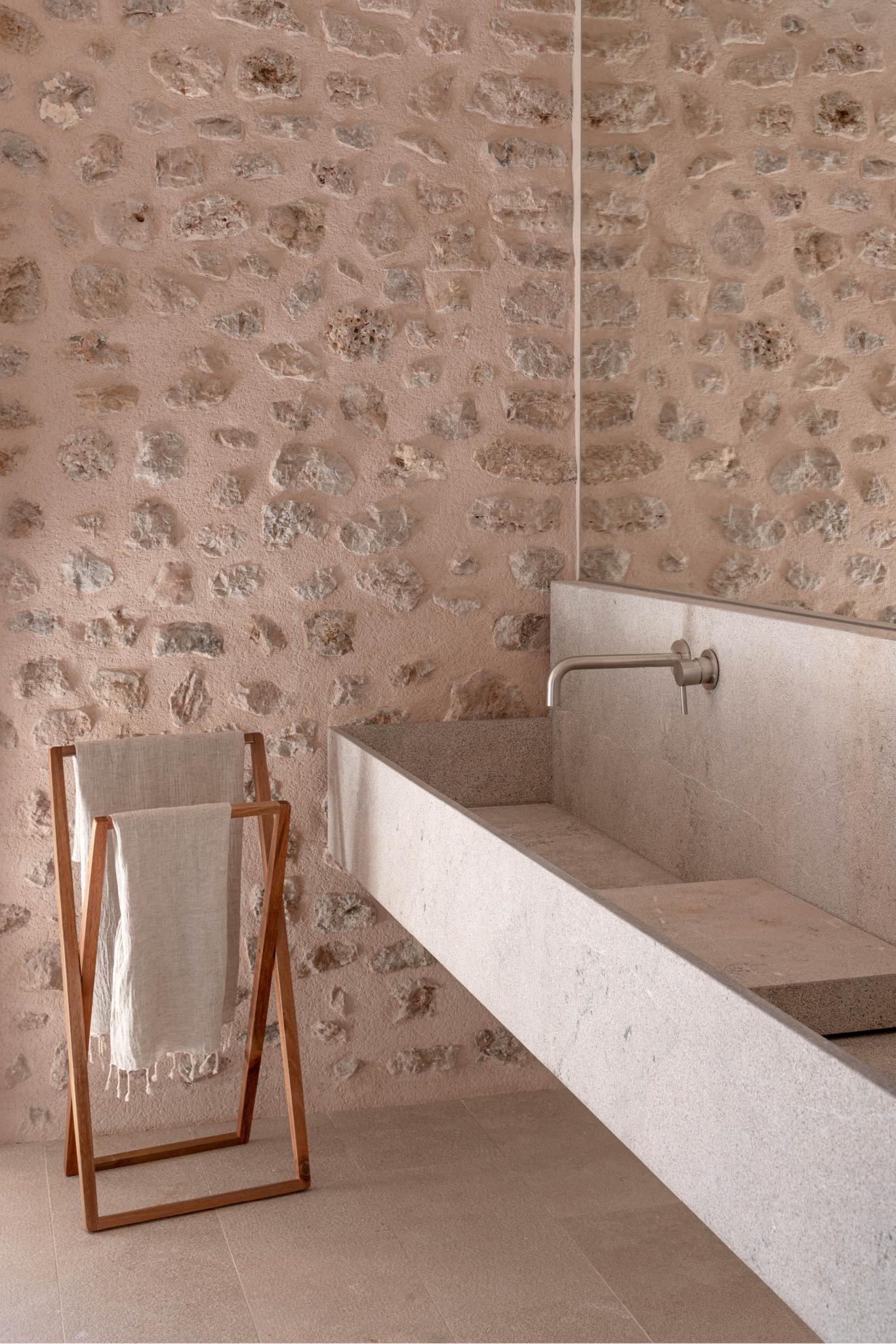 Can Gaspar, Balearic Islands — Minimalist bathroom with textured terracotta walls, concrete bathtub, wooden towel rack, and natural light through mirror.